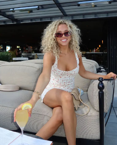 Cheerful woman with curly blonde hair and sunglasses in a white floral mini dress sitting on a beige outdoor lounge sofa at a rooftop patio, reaching to offer a lemon‑garnished cocktail.