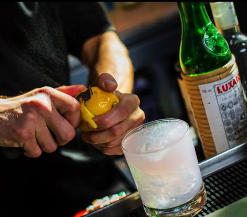 Close-up of bartender’s hands peeling a lemon twist over a frosty cocktail glass on a bar counter, with a green liqueur bottle blurred in the background.