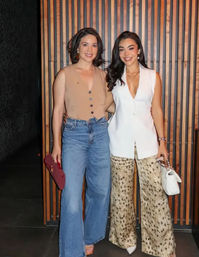 Two stylish women smiling and posing indoors against a vertical wood‑slat wall — one in wide‑leg jeans and a tan sleeveless top holding a maroon clutch, the other in a white sleeveless blazer with leopard‑print wide pants and a white handbag, casual‑chic evening look.