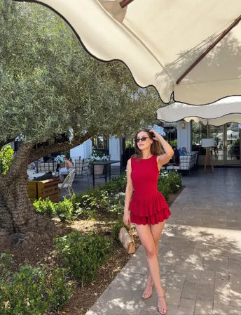 Woman in a red sleeveless ruffled mini dress, sunglasses and sandals holding a straw bag, posing under a large patio umbrella by an olive tree on a sunny outdoor café terrace.