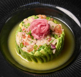 Avocado-wrapped ceviche tower of diced fish, tomato and herbs, crowned with pickled red onions and served in a bright citrus-green broth in a black bowl.