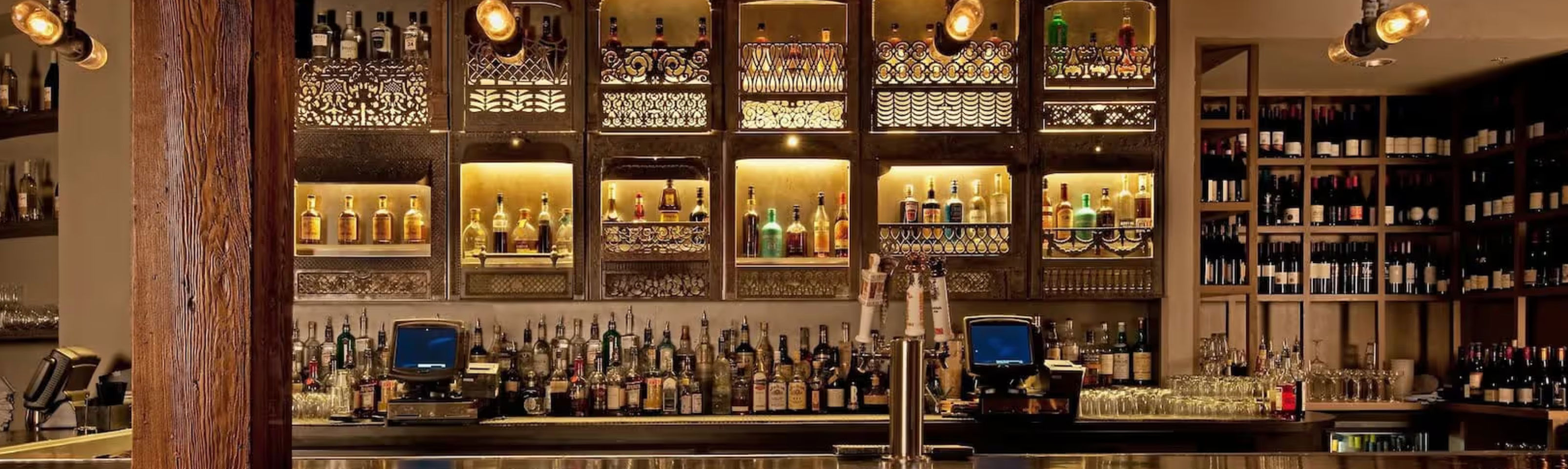 Cozy upscale cocktail bar interior with backlit wooden shelves of liquor bottles and wine racks, warm pendant lighting, exposed wooden beam, polished metal bar counter with draft taps, glassware and register stations.