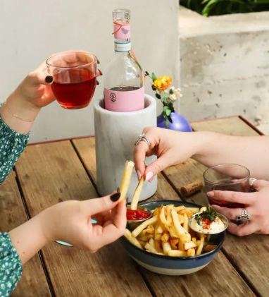 Two people sharing fries and sipping rosé on a wooden outdoor patio table — bowl of fries with ketchup and aioli, glasses of rosé, chilled rosé bottle in a marble cooler and a small vase of flowers.