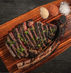 Sliced charred tomahawk (bone-in ribeye) steak, medium-rare with microgreens, on a wooden serving board with three seasoning piles on a dark tabletop