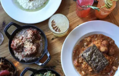 Overhead shot of vibrant shareable dishes on a wooden table: seared fish atop tomato stew, meatballs in a cast-iron skillet, creamy risotto, small sides and colorful cocktails.