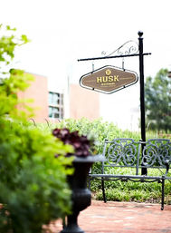 Sunny urban garden courtyard with a hanging restaurant sign, ornate wrought-iron bench on a brick patio, potted plants and lush greenery