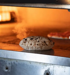 Artisan pizza dough puffing and steaming on a hot stone oven floor, charred leopard spots and glowing orange oven interior with a blurred pizza in the background.