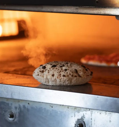 Artisan pizza dough puffing and steaming on a hot stone oven floor, charred leopard spots and glowing orange oven interior with a blurred pizza in the background.