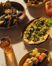 Overhead shot of a vibrant dinner spread: grilled meat and vegetable skewers, herb-topped flatbread, colorful sushi rolls with pickled ginger and wasabi, a mixed salad, and two garnished cocktails on a marble table.