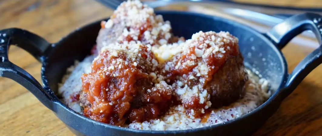 Close-up of three Italian-style meatballs in rich tomato sauce with grated cheese on creamy polenta, served in a small cast-iron skillet on a wooden table.