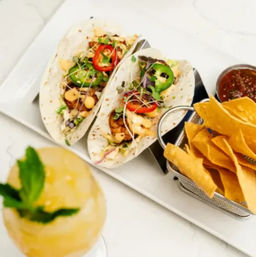 Two gourmet soft tacos with grilled filling, jalapeño slices, red pepper and microgreens, served with tortilla chips, salsa and a minty cocktail on a white marble tabletop.