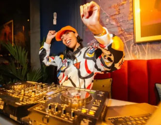 Energetic DJ in an orange cap and colorful jacket smiling while mixing on turntables at a cozy indoor lounge with red banquette and warm ambient lighting.