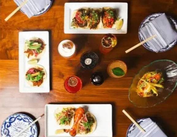 Top-down view of a wooden restaurant table spread with gourmet tacos on white plates, colorful cocktails, patterned napkins and chopsticks — lively taco-and-cocktail dining scene