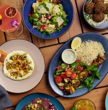 Colorful Mediterranean mezze on a wooden table — grilled skewers with rice, tomato-cucumber salad, hummus topped with pistachios, falafel and cocktails.