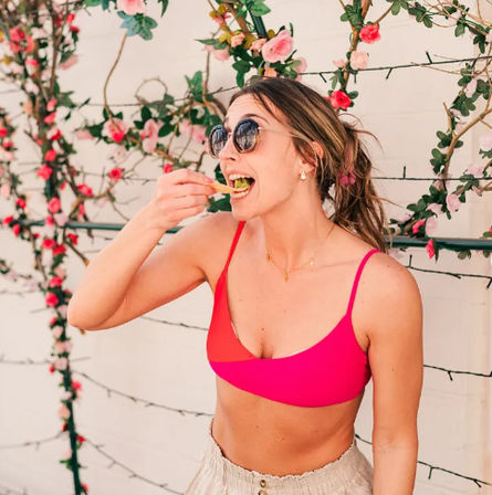 Young woman in a pink bikini top and sunglasses eating a bite outdoors in front of a flower-covered wall — summer lifestyle brunch scene