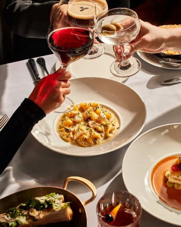 Fine-dining restaurant table with creamy gnocchi in a white bowl, hands clinking red wine and an olive martini over a white tablecloth, plated appetizers and a cocktail nearby.
