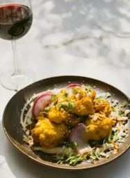 Gourmet roasted golden cauliflower florets on a brown plate with creamy sauce, sliced radishes, microgreens and toasted almond flakes, styled on a marble tabletop beside a glass of red wine.