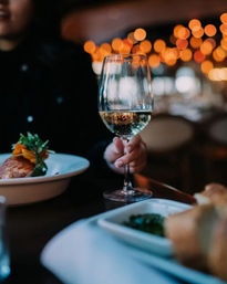 Glass of white wine held at a cozy restaurant dinner table, warm bokeh lights in the background, plated entrée and bread in the foreground.
