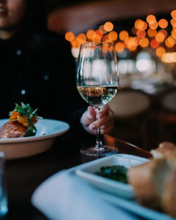 Glass of white wine held at a cozy restaurant dinner table, warm bokeh lights in the background, plated entrée and bread in the foreground.