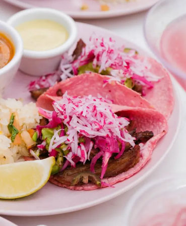 Colorful Mexican street tacos on pink corn tortillas topped with shredded radish and pickled cabbage, grilled meat, avocado salsa, rice and lime wedge with dipping sauces