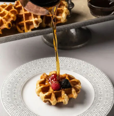 Close-up of a round Belgian waffle on a white patterned plate topped with raspberries and a blackberry as golden maple syrup is poured over it — breakfast brunch shot