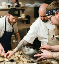 Tattooed chefs in a busy restaurant kitchen carefully plating garnished dishes, one holding a large metal mixing bowl while others arrange food into a row of bowls.