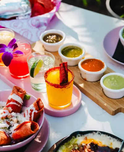 Vibrant Mexican-style spread on a sunlit patio: crispy taquitos topped with crema and cotija, colorful cocktails (mango drink with chili-rimmed glass, pink orchid garnish, lime cocktail), a wooden board of assorted green and red salsas, and bubbling queso dip.