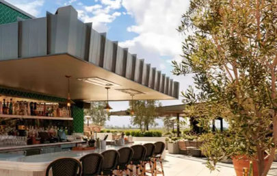 Sunlit rooftop bar patio with curved marble counter, rattan bar stools, pendant lights and stocked bottle shelves, framed by potted olive trees and a distant skyline.