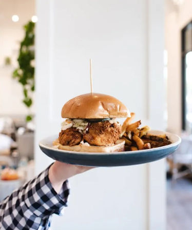 Crispy fried chicken sandwich on a brioche bun with slaw and pickles, served with fries on a blue plate held up in a bright airy café.