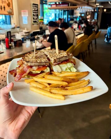 Grilled BLT on toasted sourdough with crispy bacon, lettuce and tomato, served with golden fries on a white plate in a bustling café bar.