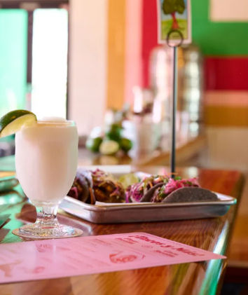 Frosty frozen margarita with a lime wedge next to a tray of colorful street-style tacos on a wooden bar in a bright casual Mexican eatery