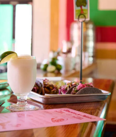 Frosty frozen margarita with a lime wedge next to a tray of colorful street-style tacos on a wooden bar in a bright casual Mexican eatery