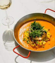 Grilled fish fillet atop seasoned yellow rice with peas and microgreens in a vibrant orange sauce, presented in a shallow pan with red handles beside a glass of white wine.