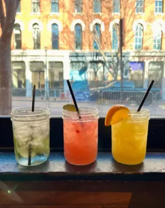 Three colorful cocktails in mason jars on a windowsill — clear lime drink, pink cocktail with lime wedge, and bright orange drink with an orange slice — blurred downtown street and brick building visible through the window.