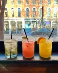 Three colorful cocktails in mason jars on a windowsill — clear lime drink, pink cocktail with lime wedge, and bright orange drink with an orange slice — blurred downtown street and brick building visible through the window.
