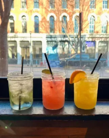 Three colorful cocktails in mason jars on a windowsill — clear lime drink, pink cocktail with lime wedge, and bright orange drink with an orange slice — blurred downtown street and brick building visible through the window.