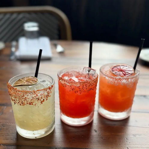 Three vibrant cocktails on a rustic wooden table — a pale margarita and two orange-red drinks in short glasses with chili-salt rims, ice, black straws and dried citrus garnishes.
