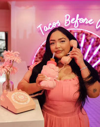 Person in a pink dress playfully holding a pink retro telephone handset to their ear while sipping from a matching novelty pink drink bottle, standing at a pink-themed counter with a vase of pink flowers and a neon cursive sign in the background.