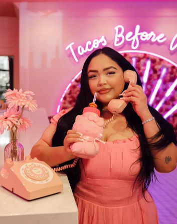 Person in a pink dress playfully holding a pink retro telephone handset to their ear while sipping from a matching novelty pink drink bottle, standing at a pink-themed counter with a vase of pink flowers and a neon cursive sign in the background.