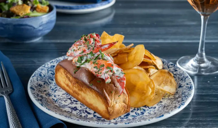 Buttery toasted brioche lobster roll topped with chilled lobster salad and herbs, served with crisp kettle-style potato chips on a blue patterned plate on a dark wood table with a wine glass nearby.