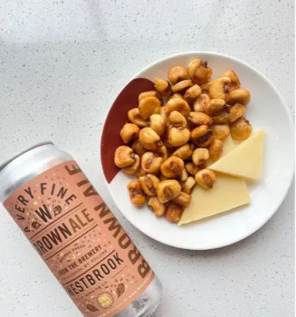 Can of brown ale beside a white plate of crunchy toasted corn nuts and two cheese wedges on a speckled kitchen countertop — a casual beer and snack pairing.
