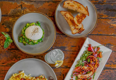 Overhead shot of a rustic wooden table with a colorful Mediterranean brunch spread: burrata on pea salad, toasted bread slices, grilled octopus salad with chickpeas and pickled onions, pasta, and two refreshing drinks.