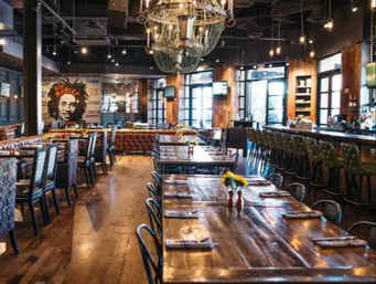 Rustic-modern restaurant interior with long wooden communal tables, leather banquette seating, industrial chandelier, bar with stools, exposed brick mural and small sunflower centerpieces, cozy urban gastropub vibe.