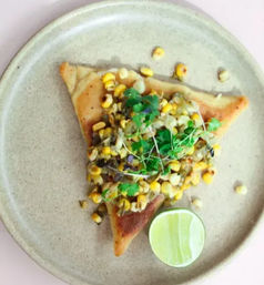 Golden triangular toast topped with charred corn salad, cilantro microgreens and a lime half on a beige ceramic plate