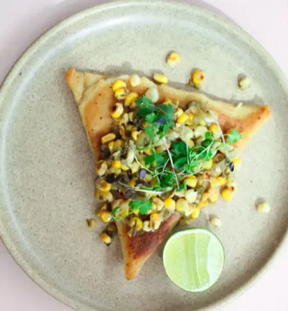 Golden triangular toast topped with charred corn salad, cilantro microgreens and a lime half on a beige ceramic plate