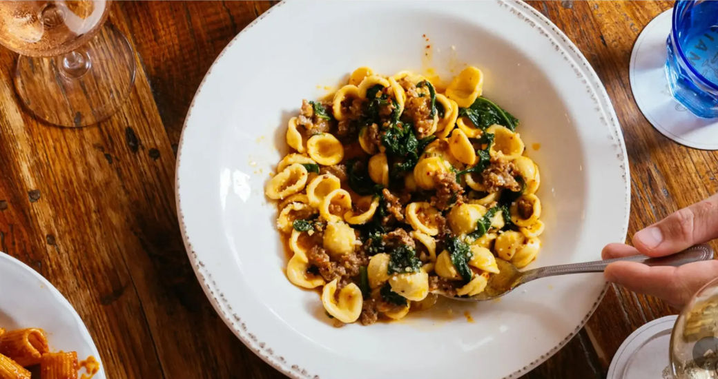 Orecchiette pasta with Italian sausage, wilted spinach and tomato sauce in a white bowl on a rustic wooden table, fork poised to scoop