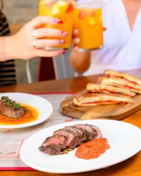 Sliced medium-rare steak with red pepper sauce on a white plate, panini on a wooden board, and two hands clinking orange fruit cocktails at a restaurant table.