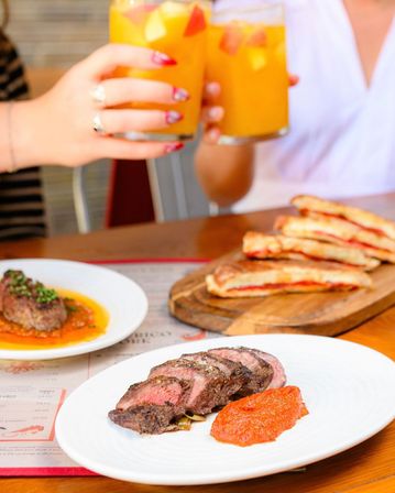 Sliced medium-rare steak with red pepper sauce on a white plate, panini on a wooden board, and two hands clinking orange fruit cocktails at a restaurant table.