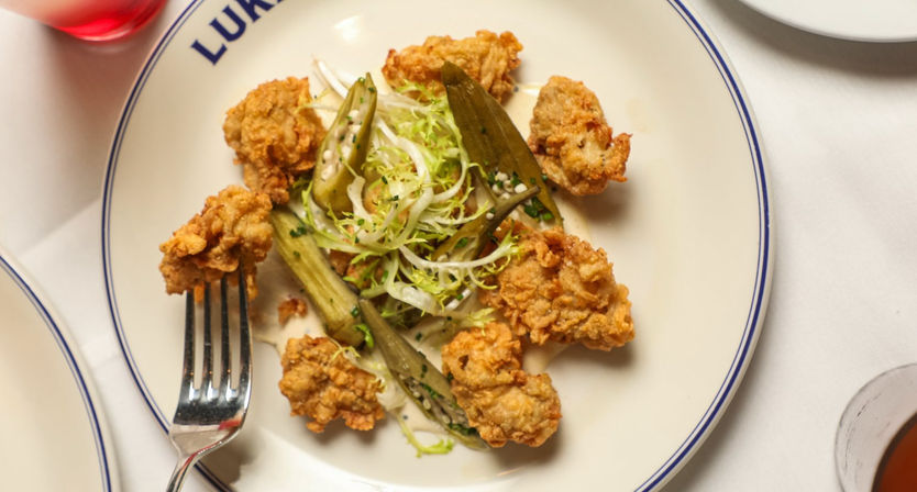 Golden-brown crispy fried oyster bites with pickled okra and frisée salad arranged on a white blue-rimmed plate, fork and drink visible on a restaurant table