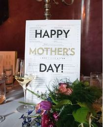 Festive Mother's Day sign reading "HAPPY MOTHER'S DAY!" standing on a restaurant table beside a white wine glass and a colorful floral centerpiece.